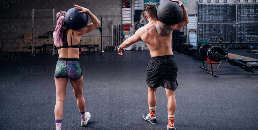 Young woman and man training together, carrying atlas ball on shoulders in gym, rear view